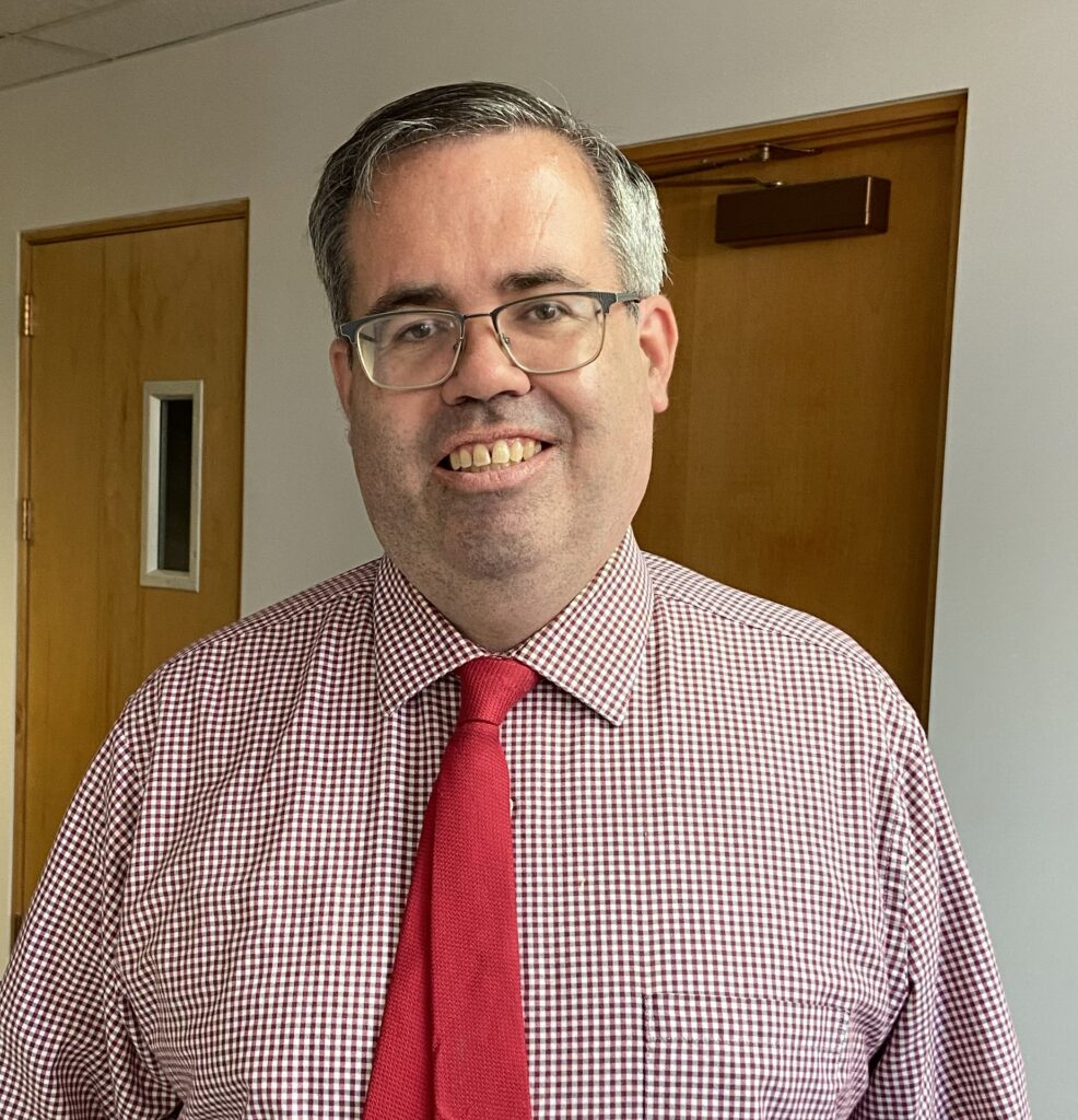 Headshot of Michael Courtney, smiling, in a red checked shirt and red tie, standing in front of two doors.